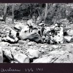 A photograph c. 22 June 1911, of a group of 17 people and 2 dogs on rocky vantage points in an almost dry Araluen creek bed.
